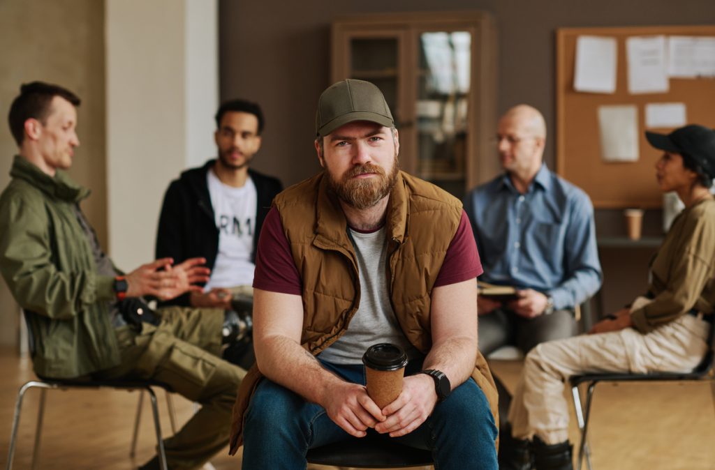 bearded man with cup of coffee looking at camera against group of people